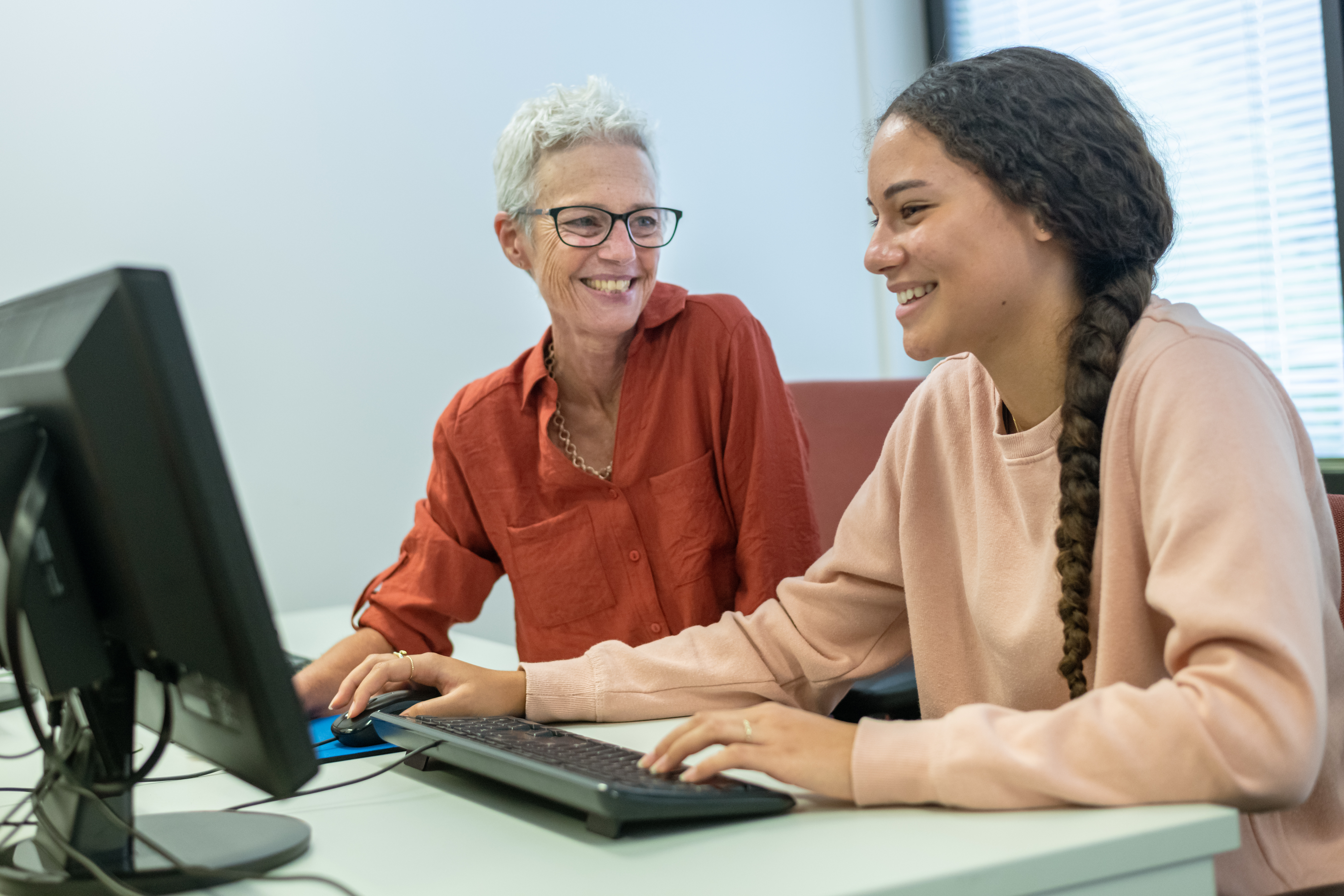 A teenage girl smiles while working on a computer in the school library with the help of an adult female teacher or tutor.